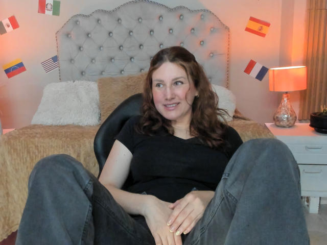 KatyaRogersX sits on a black chair, legs spread, wearing a black shirt and gray pants, smiling with brown wavy hair, hands on her pussy, in a bedroom.