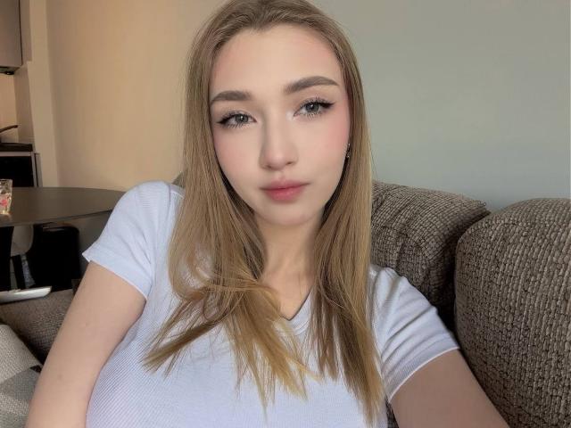 DenisseReins hat langes, blondes Haar, ein weißes T-Shirt und dezentes Make-up mit rosafarbenem Lippenstift. Sie sitzt auf einem braunen Sofa und blickt mit einem ruhigen Ausdruck direkt in die Kamera..