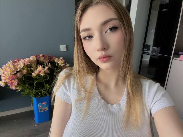 DenisseReins a la peau claire, les cheveux blonds, et elle porte un t-shirt blanc. Elle regarde l'appareil photo avec une légère moue, et il y a un vase en forme de fleur bleue en arrière-plan..
