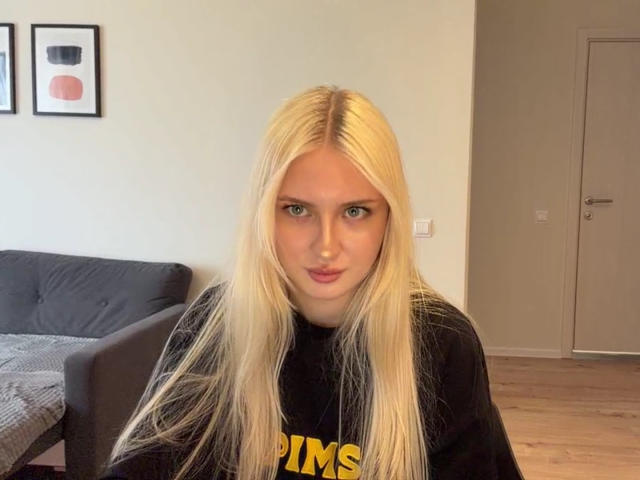 NancyBijou tem cabelos loiros longos, olhos azuis e uma expressão emburrada. Ela está vestindo uma camisa preta com texto amarelo e está em uma sala com um sofá cinza..