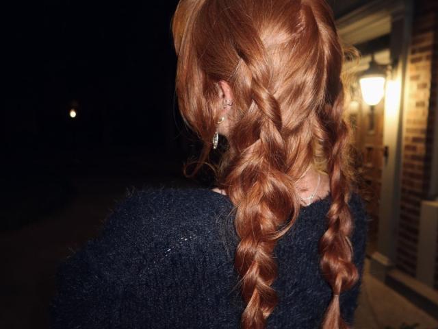 BariBenafield mit roten, geflochtenen Haaren, trägt einen schwarzen, flauschigen Pullover, Ohrringe sind sichtbar, mit dem Rücken zur Kamera, nachts, draußen, beleuchtet von einer Lampe auf der rechten Seite..