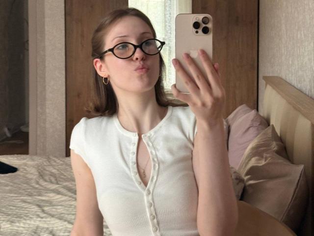 KenaCly avec des lunettes, les lèvres pincées, une chemise blanche à boutons, montrant ses seins, tenant un téléphone, dans une chambre, prenant un selfie. Cheveux blonds, peau claire, petits boucles d'oreilles, lit en bois, oreillers..
