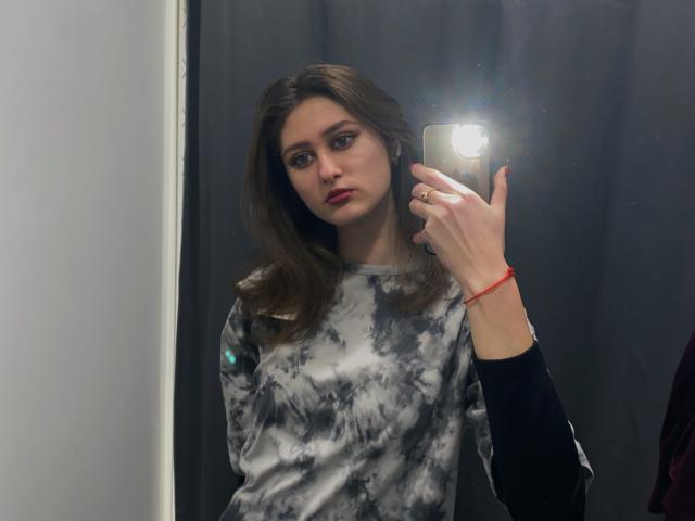 HangBielefield, com cabelo castanho ondulado, batom vermelho e uma camisa cinza tie-dye, tirando uma selfie no espelho com o flash, usando uma camiseta preta de manga comprida por baixo..