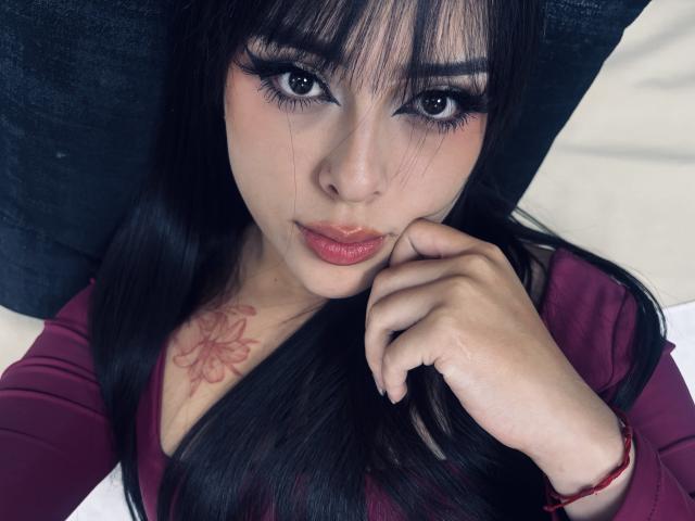 AlisonTatcher with black hair, full eyebrows, and thick eyelashes, resting her chin on her hand, wearing a maroon top, tattoo on her chest, looking directly at the camera.