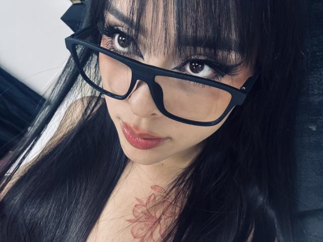 AlisonTatcher with dark hair, glasses, red lipstick, and a tattoo on her chest, staring into the camera with a slight smile. Boobs are partially visible.