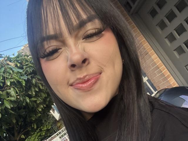AlisonTatcher's face, showing glossy pink lips, slightly upturned nose, and long black eyelashes, with straight black hair, and a subtle smile, in a bright outdoor setting.