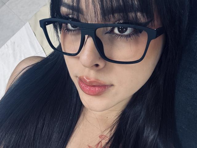 AlisonTatcher has dark, straight hair, black-framed glasses, and shiny pink lips. She's looking at the camera with a slight smile, showing off her cute, sexy face.