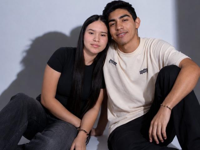 CloeAndJacob, una chica asiática atractiva con cabello negro largo, senos pequeños y una camisa negra, está sentada al lado de Jacob, un hombre latino sexy con bronceado, una camiseta blanca y pantalones negros..