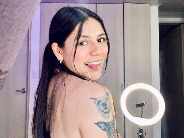 SophiaMarx with long black hair, smiling with tongue out, showing tattoo on her left shoulder, standing near a ring light, topless, showing her bare back and small earrings.