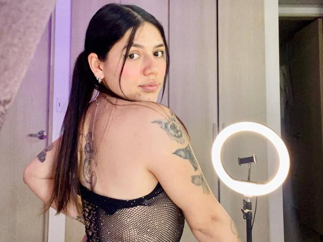 SophiaMarx with long black pigtails, looking over her shoulder, wearing a black mesh top, showing tattoos on her right arm and back, standing near a ring light.