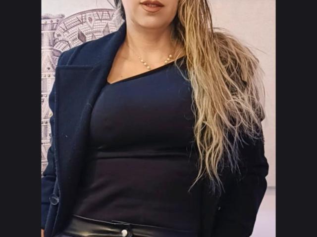 DianaPearlsMilf con lunghi capelli biondi, una maglietta nera a V e una giacca nera, che mostra il seno, indossa pantaloni di pelle nera e una collana d'oro, labbra carnose..