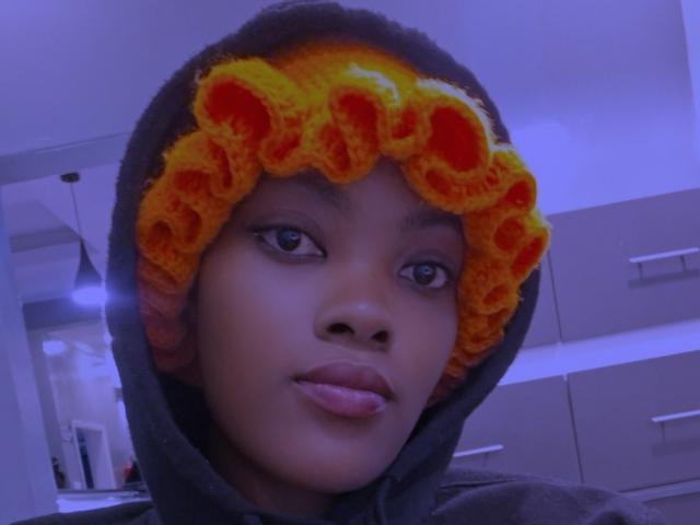 ThickianaSexy a la peau brun foncé, porte un bandeau en crochet jaune et orange avec des poches orange, ainsi qu'une capuche grise, avec une légère moue et un visage illuminé de bleu..