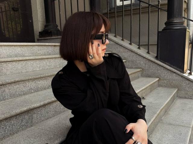 Elina Haycock mit einem Bob-Haarschnitt, schwarzen Sonnenbrillen, einem schwarzen Kleid und einer schwarzen Hose, sitzt auf grauen Steinstufen, hält ihr Telefon ans Ohr und trägt einen Ring und Ohrringe..