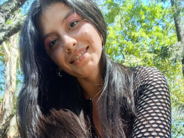 MadissonFontaine with long black hair, wearing a black zip-up top and fishnet sleeves, showing her big tits and smiling down. She's standing in a sunny forest.