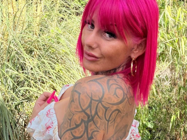 Pinki-Inked: Mit leuchtend rosa Haaren, Tattoos an der rechten Schulter, goldenen Creolen und einem Nasenring, lächelt sie in die Kamera und zeigt ihre Brüste in einem Blumenkleid..