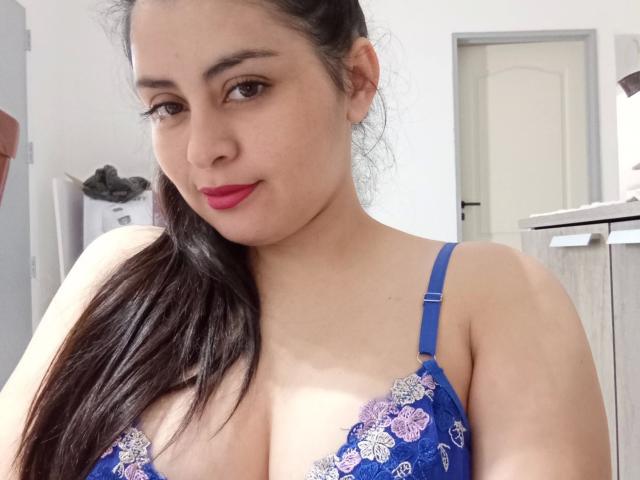 VeliSex – eine Frau mit dunklem Haar im Pferdeschwanz, die ein blaues Spitzen-BH mit violetten Blumen trägt, ihre Brüste zeigt, roten Lippenstift trägt und ein leichtes Lächeln hat, aufgenommen in einem Badezimmer..
