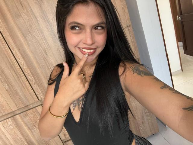 NyraVanel mit langen, schwarzen Haaren, lächelnd, mit einem Finger am Mund, schwarzes Tanktop, sichtbare Tattoos am Arm und an der Brust, goldenes Armband, braune Holztür im Hintergrund..