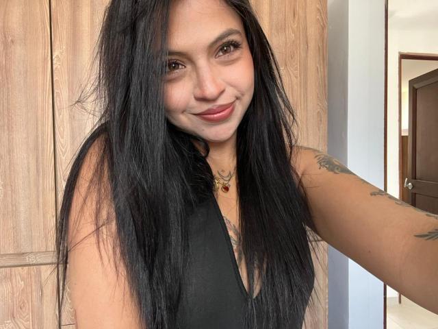 NyraVanel, com cabelos longos e pretos, sorrindo, vestindo uma blusa preta sem mangas, mostrando tatuagens em seu braço direito, e um colar com uma pedra vermelha, em um selfie..