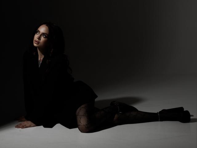 VikiPatti has dark hair, a black outfit, and patterned black tights, lying on her side with her ass up, in a dimly lit, shadowy setting.