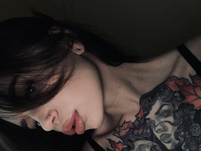 VikiPatti, avec ses cheveux brun foncé, ses lèvres roses et pulpeuses, et un anneau de nez, exhibe un tatouage noir et rouge détaillé sur son épaule. Photo sombre et mélancolique..