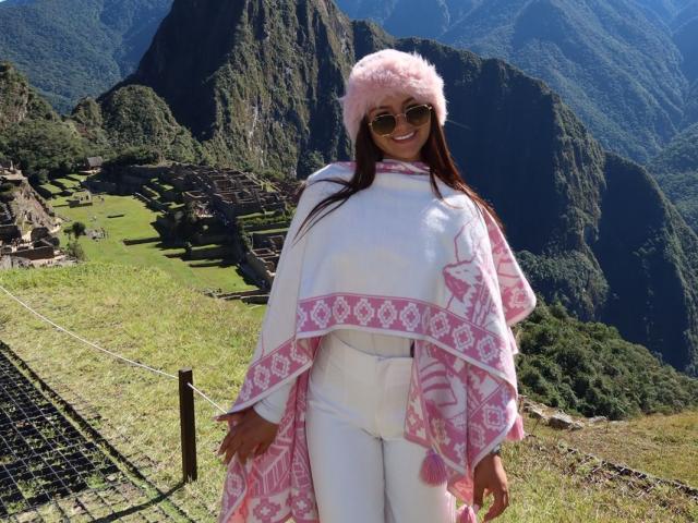AlessiaFerry with pink furry hat, white pants, and pink patterned shawl, smiling, standing in sun, wearing dark sunglasses, with green mountains and ruins in background.
