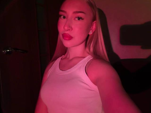 Joette Shigeta, blonde avec du rouge à lèvres, portant un débardeur blanc, éclairage rose, de petites boucles d'oreilles et un piercing au nez, se prend en photo dans une pièce peu éclairée..