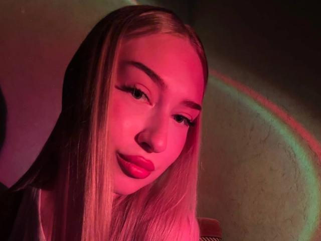 Het gezicht van Joette Shigeta is verlicht met roze en rode licht, en je ziet lang, recht blond haar, opvallende wenkbrauwen en roze lippenstift, met een ontspannen en licht gekanteld hoofd..