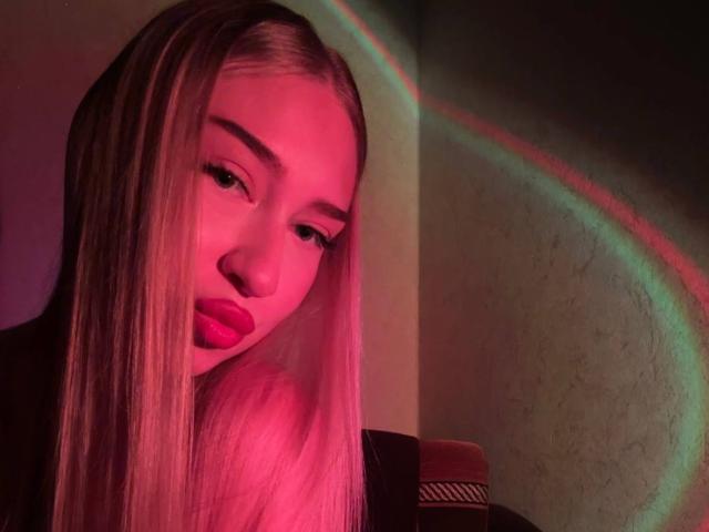 Joette Shigeta, met lang, steil blond haar, roze-rode verlichting en een lichte frons, haar roze lippenstift die overeenkomt met de neonregenboogreflectie op de muur..