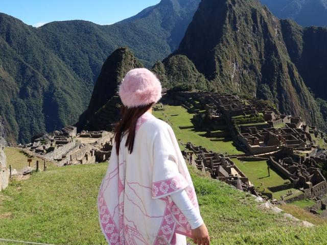 AlessiaFerry in a pink fluffy hat, white robe with pink patterns, standing on grassy hill, facing ancient ruins with green mountains in background, clear blue sky above.
