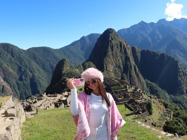 AlessiaFerry with pink fluffy hat, white shirt, and pink jacket, holding phone, standing in front of Machu Picchu, mountains and ruins in background, sunny day, blue sky.
