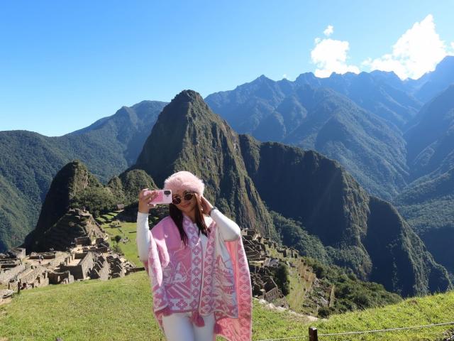 AlessiaFerry with pink hat, white patterned pink coat, and sunglasses, holding pink sunglasses, standing in green mountain landscape with ruins in background, clear blue sky above.