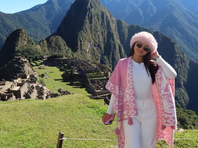 AlessiaFerry with pink fuzzy hat, pink scarf, white clothes, and sunglasses, standing in front of Machu Picchu, holding phone, smiling, green mountain background, clear blue sky.