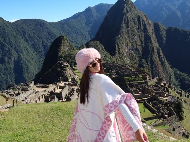 AlessiaFerry with pink fluffy hat, white robe with pink patterns, and sunglasses, standing on grassy hill, looking back over her shoulder, with ancient ruins and mountains behind.