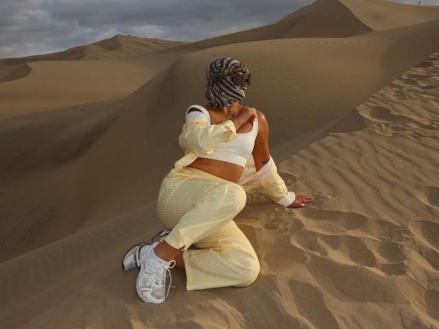 AlessiaFerry, met donkere huid, in een witte crop top en geel geruite broek, knielend in het zand, haar borsten aanraakend, met witte sneakers en een gestreepte hoofddoek..