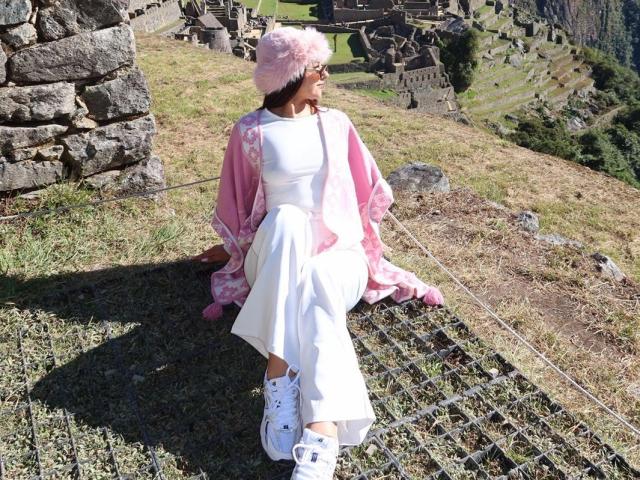 AlessiaFerry with pink fluffy hat, white dress, and pink robe, sitting on grass, white sneakers, looking to the side, stone wall in background, sunny day, grassy hill.