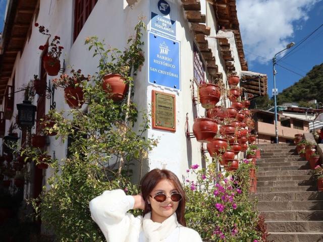 AlessiaFerry with brown hair, wearing sunglasses and a white turtleneck, stands in front of a white building with red pots of flowers, smiling with her hand on her head.