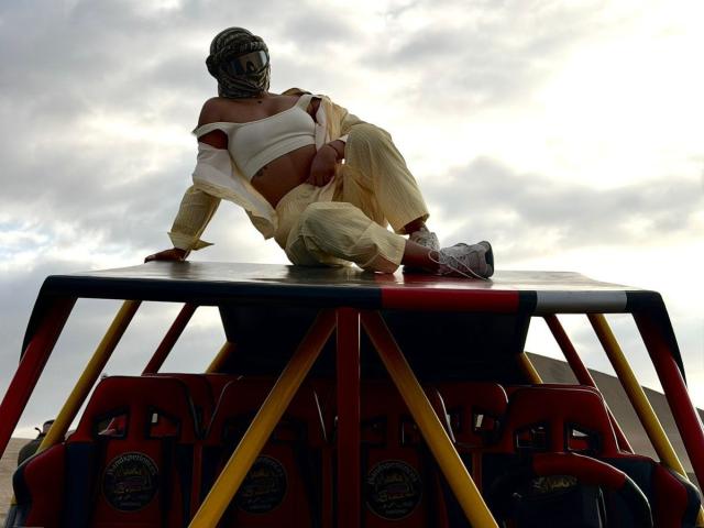 AlessiaFerry, met een gebruinde huid, draagt een masker, een witte top met open schouder en gele broek, en ligt ontspannen op het dak van een rode en gele vrachtwagen, waarbij haar achterwerk zichtbaar is..