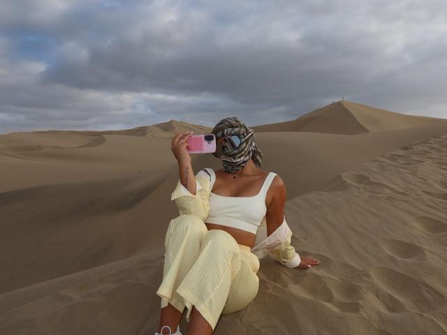 AlessiaFerry with tan skin, white crop top, yellow pinstriped pants, and patterned headscarf, sitting in sand, holding pink phone, showing tattooed arm, under cloudy sky.