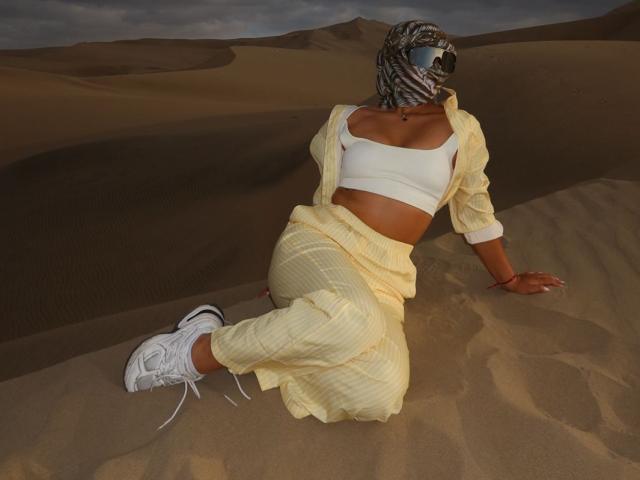 AlessiaFerry, met een bruine huid, een witte sportbeha, een geel geruit shirt, een bijpassende rok, witte sneakers en een bruin gestreept masker, zittend in een woestijn met zandduinen..