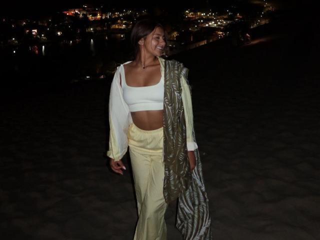 AlessiaFerry, met een bruine huid, bruin haar, kleine borsten, gekleed in een witte crop top, gele broek en een groene sjaal met een patroon, lachend 's nachts op een donker strand..