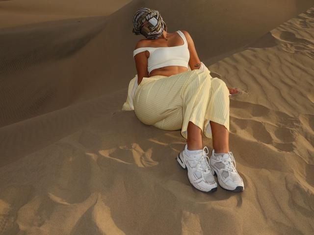 AlessiaFerry sits in sand with her back against a dune, wearing a white crop top, yellow skirt, and white sneakers. She has a headscarf and visible ass.