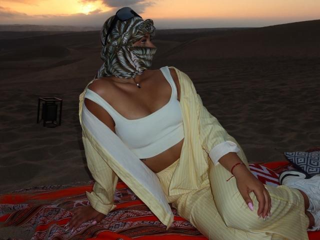 AlessiaFerry, usando uma máscara estampada, um cropped branco, calças amarelas com listras finas e um casaco, sentada em um cobertor vermelho em um deserto ao pôr do sol, mostrando seus seios..