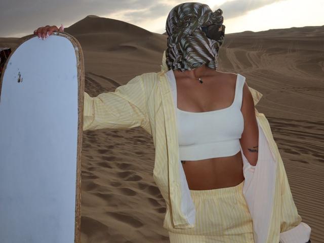 AlessiaFerry with a patterned scarf, yellow striped outfit, white sports bra, and matching pants, holding a snowboard, standing in a desert, showcasing her small tits and toned ass.