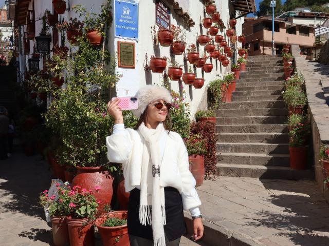 AlessiaFerry with fluffy white hair, wearing a white sweater, black skirt, and pink sunglasses, holding a pink phone, standing in front of potted plants on a sunlit staircase.