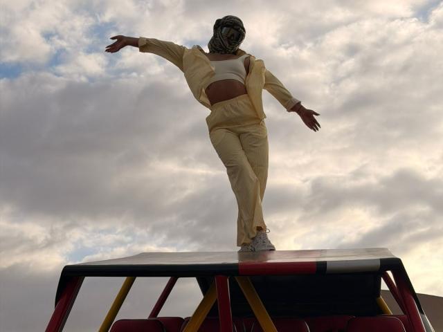 AlessiaFerry stands on a red and yellow structure, arms out, wearing a beige outfit, white crop top, and headscarf, with white sneakers, under a cloudy sky.