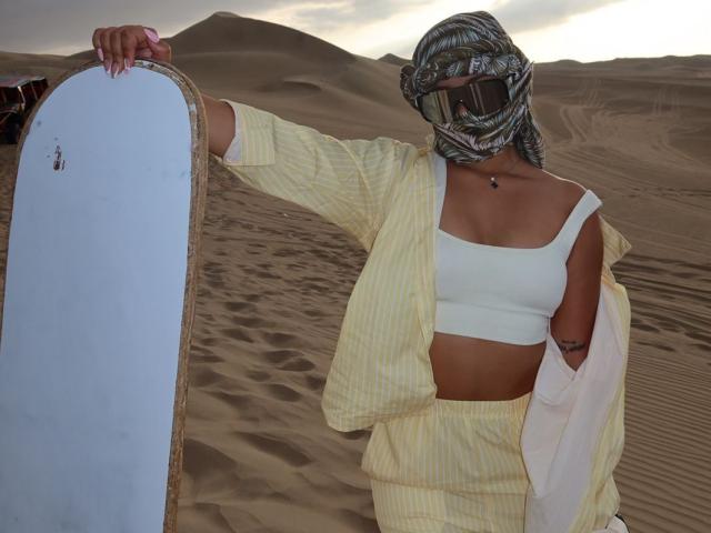 AlessiaFerry staat in een woestijn, rechtopstaand met een snowboard, gekleed in een geel gestreept outfit, een witte sportbeha en een patroonmasker, waarbij haar borsten zichtbaar zijn en ze een klein achterwerk heeft..