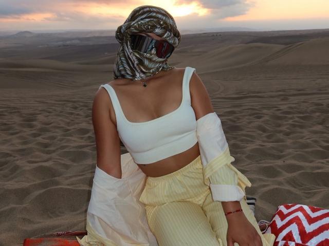 AlessiaFerry, com seios médios, vestindo uma cropped branca, calças amarelas e uma máscara com padrão prateado, sentada em um deserto arenoso ao pôr do sol, com uma camisa branca fora..