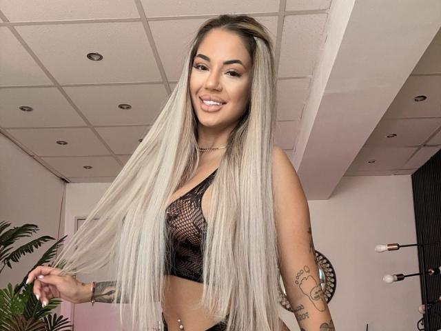 Yukenzi, con lunghi capelli biondi e lisci, indossa una maglietta nera in rete che lascia intravedere i suoi piccoli seni, sorride, ha tatuaggi sul braccio destro e un piercing all'ombelico..