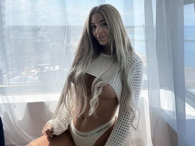 Yuukenzi, con lunghi capelli biondi, indossa una maglietta bianca in rete che rivela il seno e biancheria intima Calvin Klein bianca, seduta di fronte a una finestra illuminata dal sole..