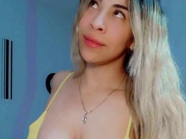 DayanaPrecious has blonde hair, light skin, and pink lips. She's wearing a yellow top, showing her tits, and a silver necklace with a small pendant. She's looking sideways.